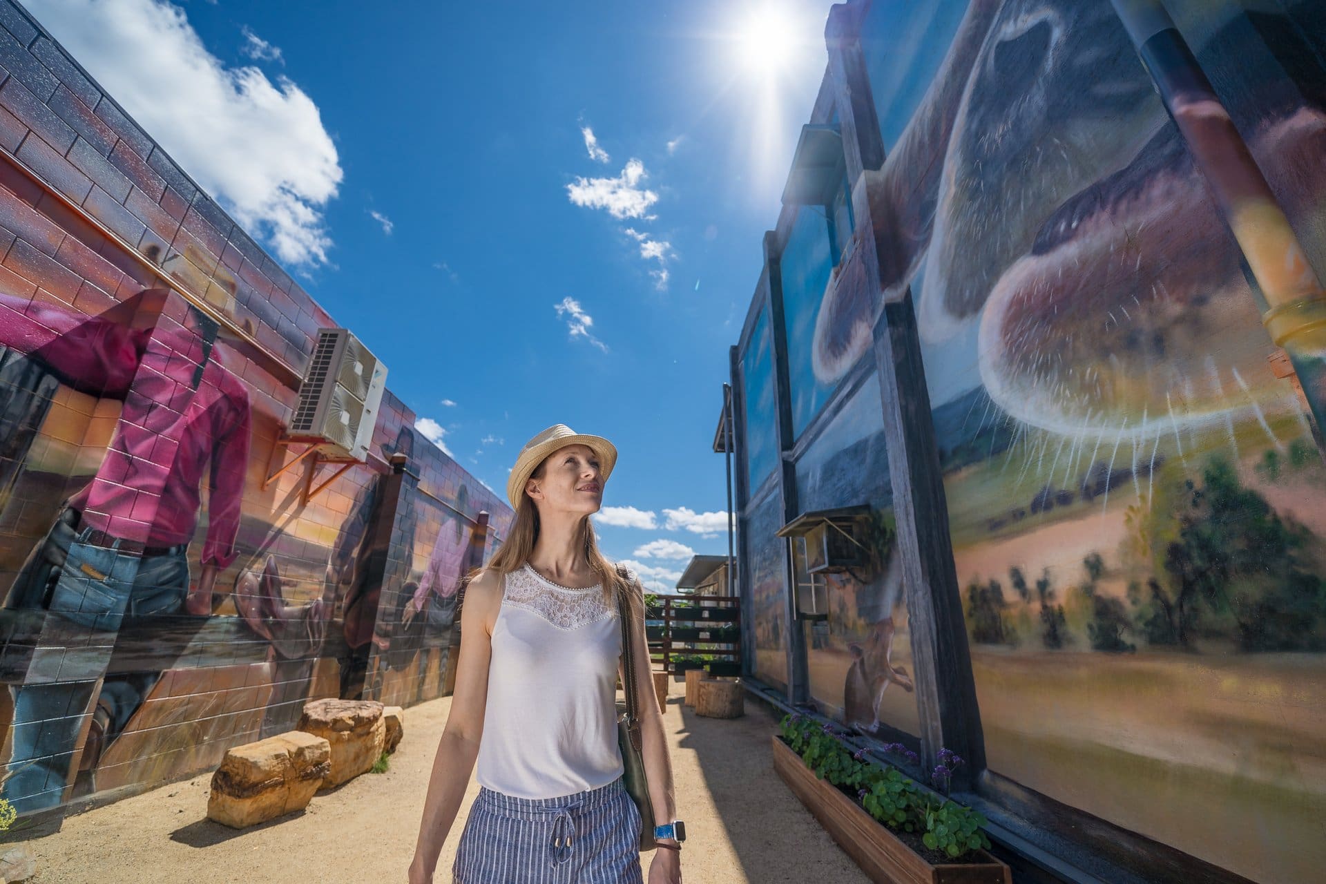 Visitor walking through a narrow outdoor laneway lined with large, colourful murals depicting local landscapes and wildlife, under a bright blue sky with sunlight overhead.