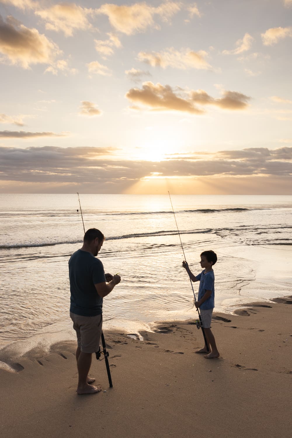 Two people beach fishing at sunrise on a calm coastline, standing on wet sand with rods as gentle waves roll in.
