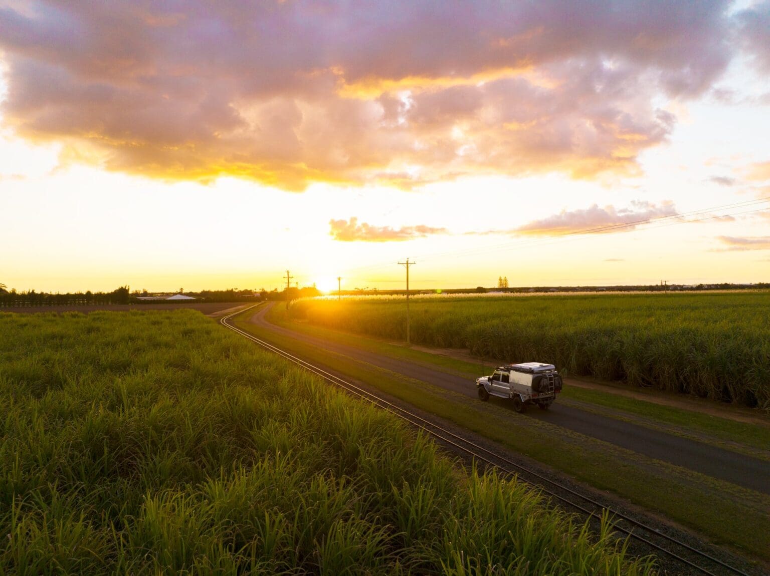 A four-wheel-drive vehicle travels along a rural road beside railway tracks, surrounded by sugar cane fields at sunset.