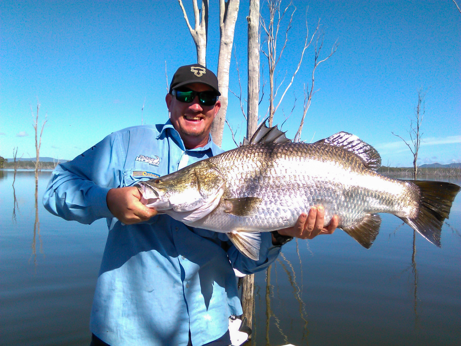 Angler holding a large barramundi at Lake Monduran in the Bundaberg region, with still water and tree trunks in the background.